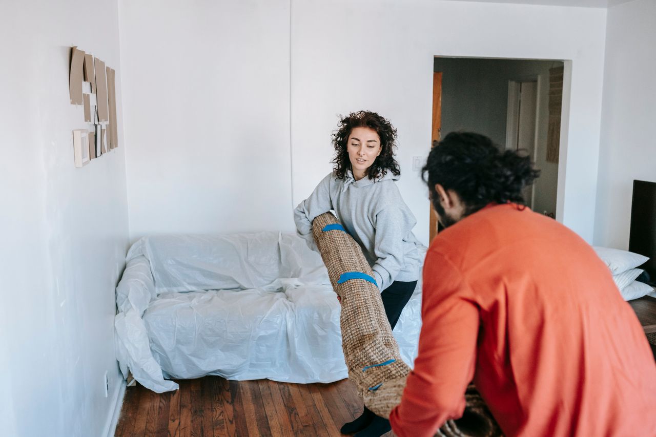 A couple lifting a rolled carpet in a minimally furnished apartment living room.